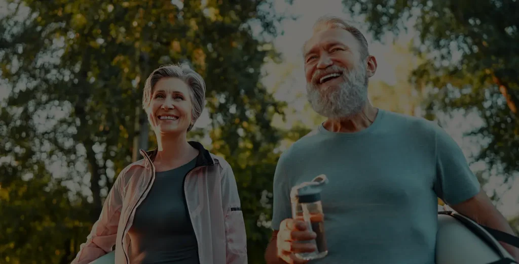 Smiling older woman and man in athletic wear stand outdoors in a park, holding water bottles and yoga mats, with sunlight filtering through green trees in the background.