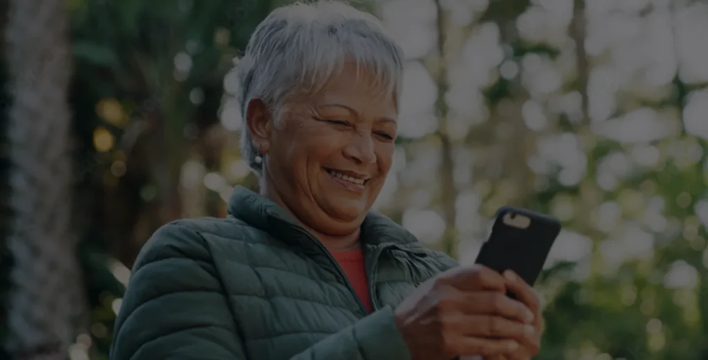 An older woman with short gray hair, wearing a green jacket, smiles as she looks at her smartphone outdoors. Sunlight and blurred trees are visible in the background.