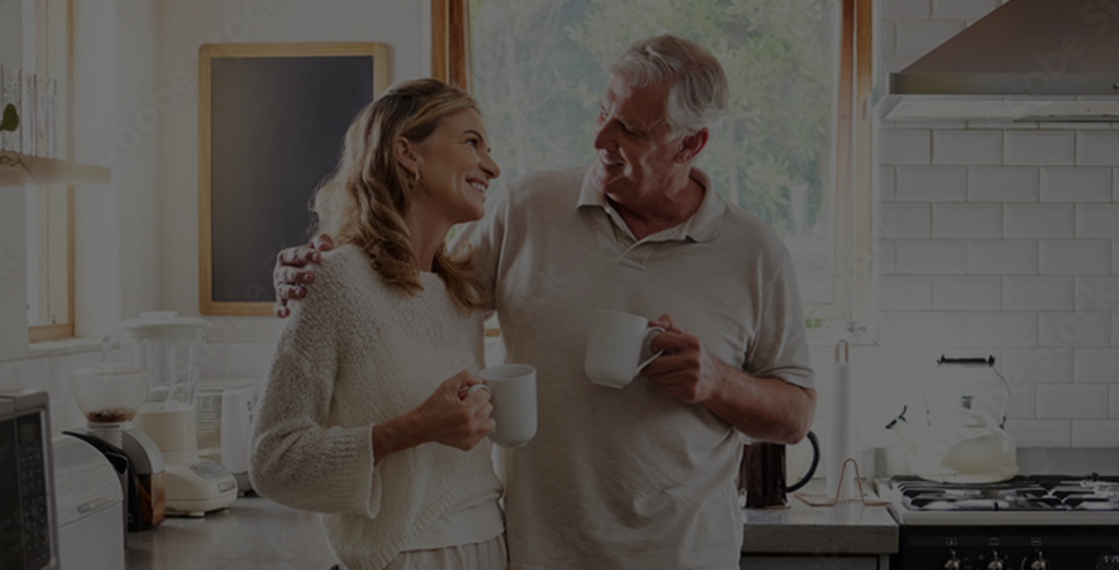 An older couple stands in a bright kitchen, smiling at each other and holding mugs. The man has his arm around the woman as they enjoy a warm, relaxed moment together.