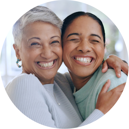 Two women smiling and hugging each other closely, both looking happy and joyful. One has short gray hair and the other has long dark hair tied back. They appear to be enjoying a warm, affectionate moment together.