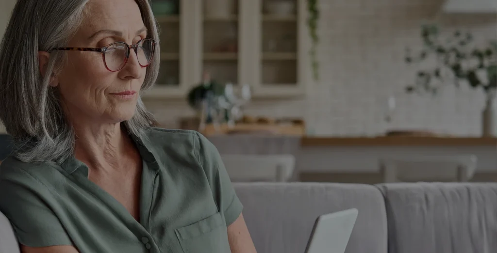 An older woman with gray hair and glasses sits on a sofa, looking at a tablet. She wears a green blouse, and a kitchen with shelves and plants is blurred in the background.