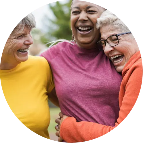 Three older adults stand close together outdoors, smiling and laughing warmly. They appear joyful and embrace each other, dressed in casual colorful shirts. The background is slightly blurred greenery.