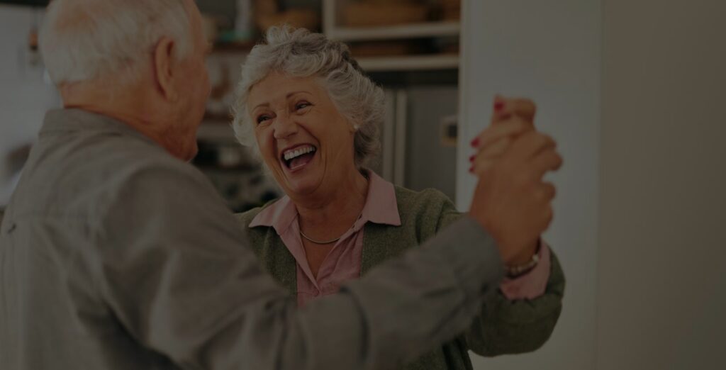 An elderly couple smiles and holds hands while dancing together indoors, enjoying a joyful moment.