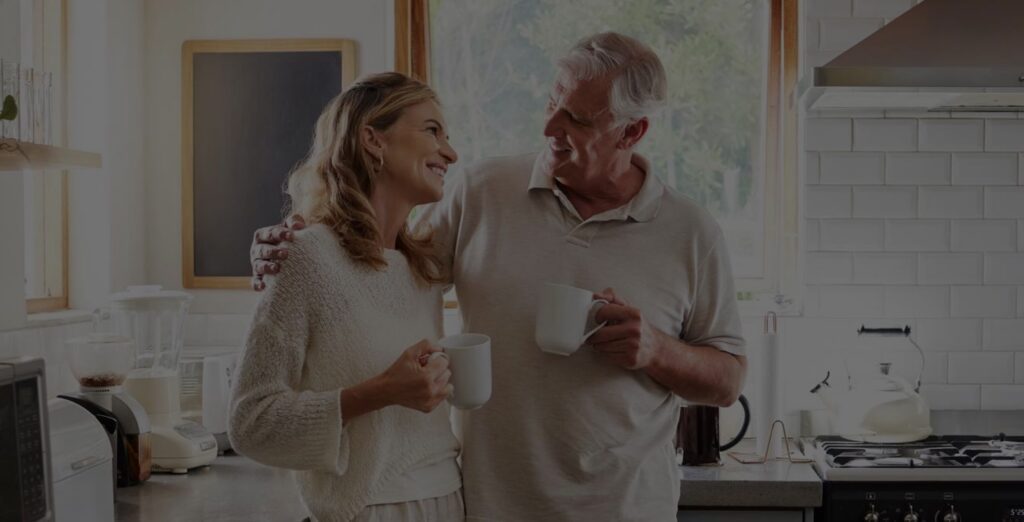 An older couple stands in a bright kitchen, smiling at each other while holding coffee mugs. The man has his arm around the woman, and sunlight streams through the window behind them.