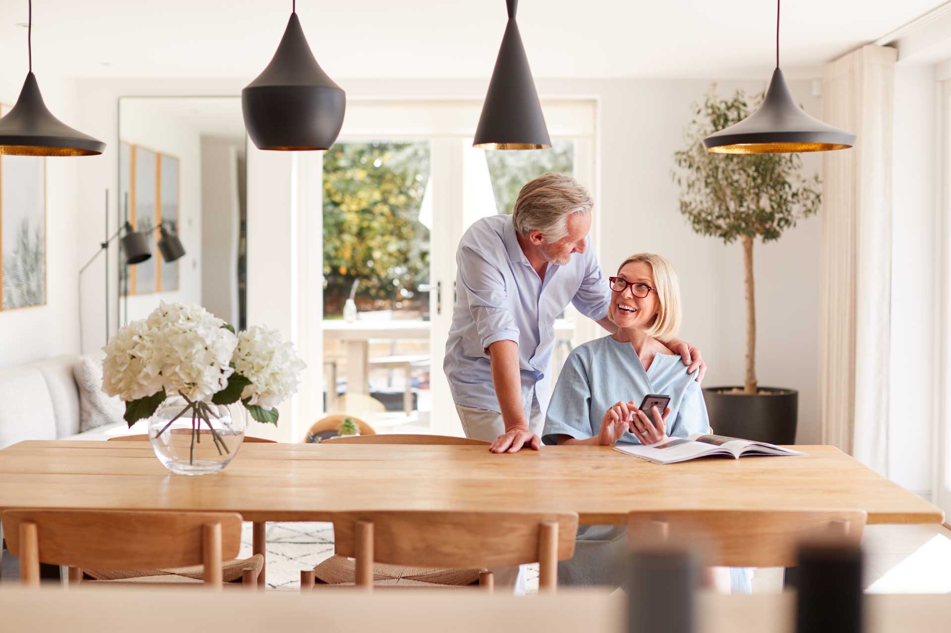 A smiling older couple in a modern, bright dining room; the woman sits at a wooden table with a phone and magazine, while the man stands behind her, resting his arm on her shoulder.